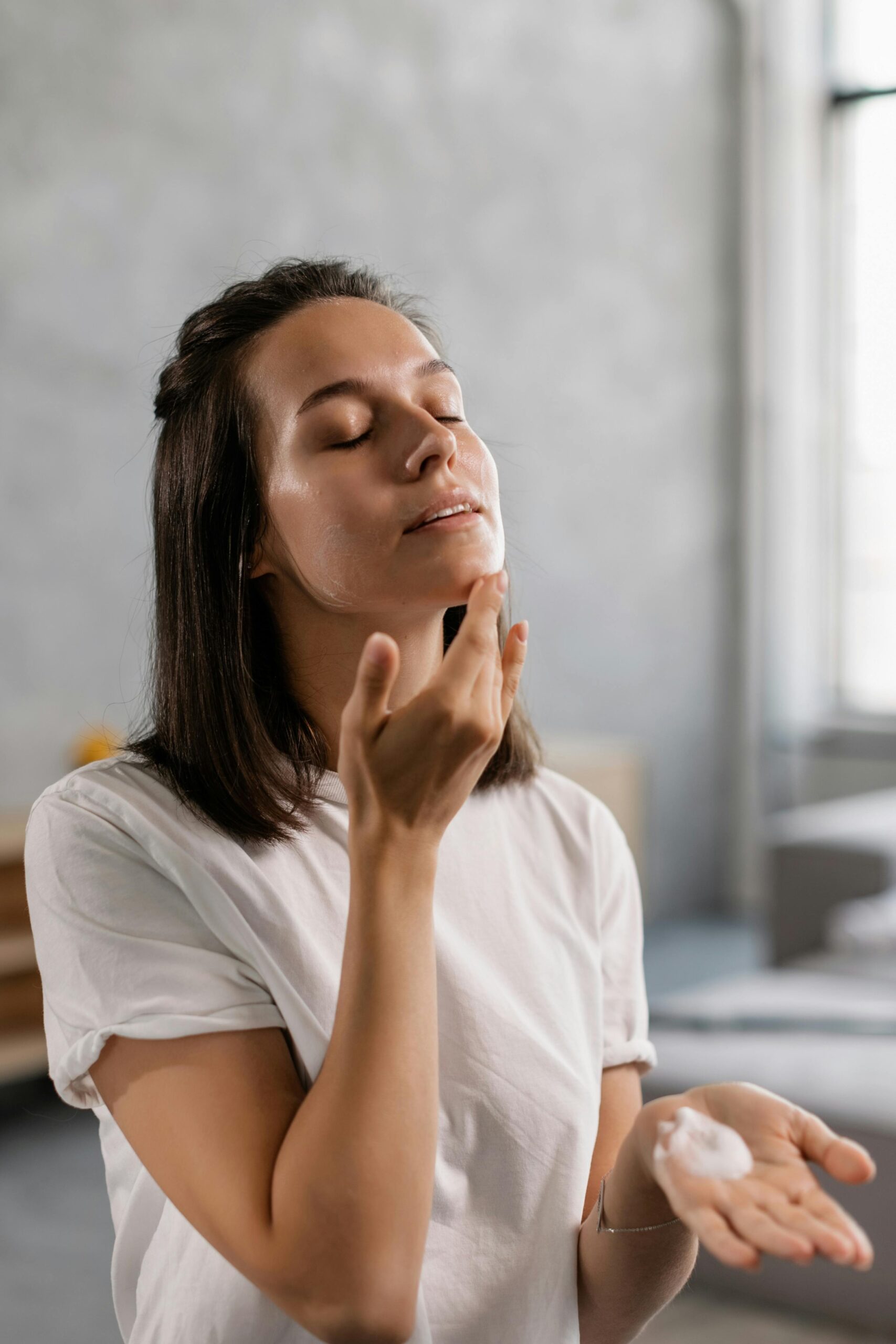 Young woman applying skincare cream to her face in a bright indoor setting.